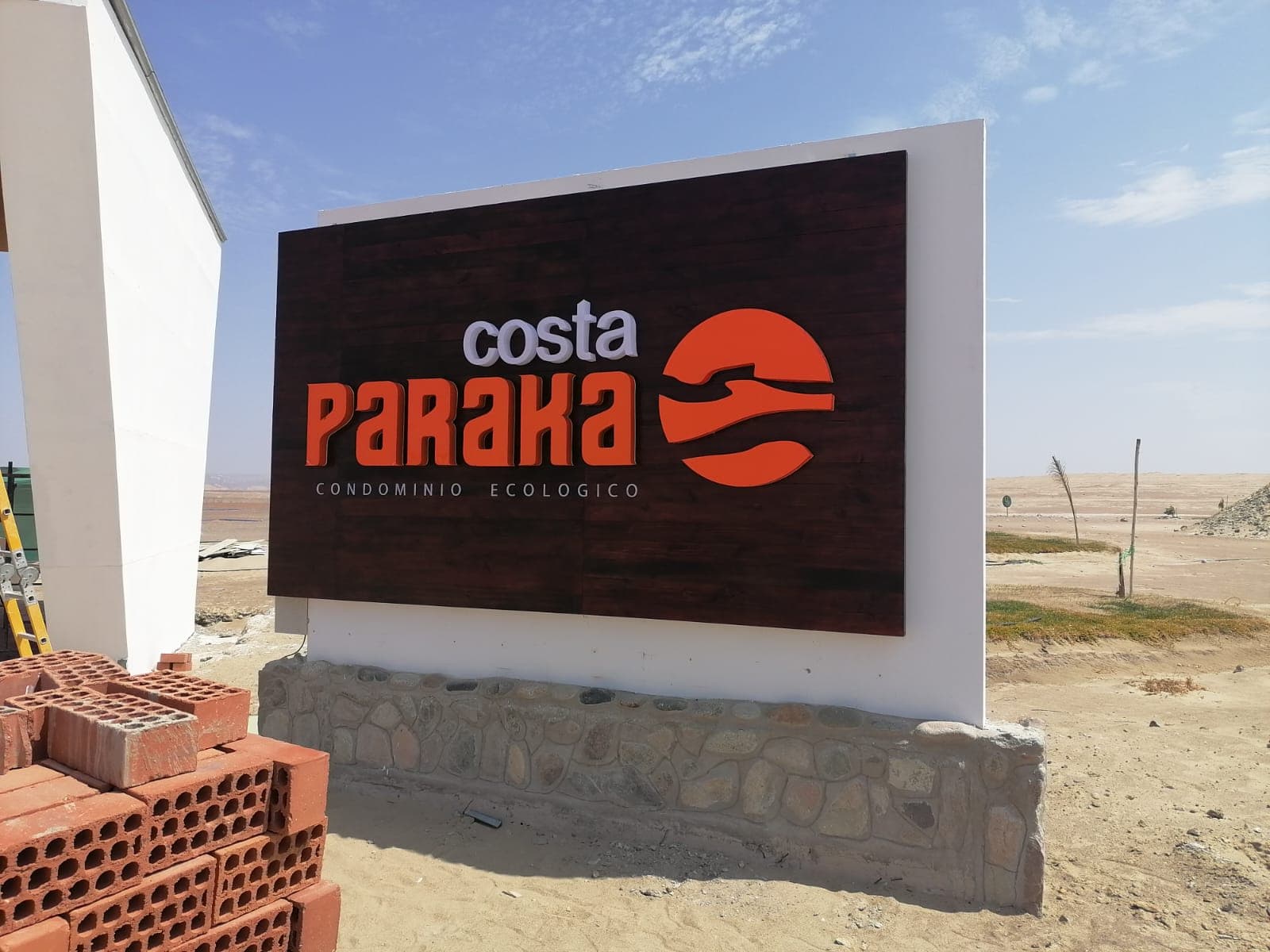 Señal exterior Costa Paraka con letras naranjas y blancas sobre madera oscura